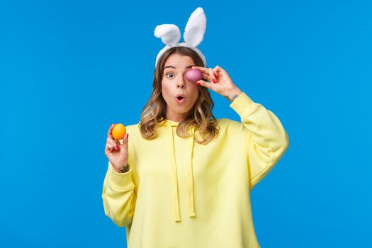 Holidays, traditions and celebration concept. Amused lovely european female blonde in rabbit ears, look excited and surprised as holding two painted Easter day eggs, blue background.