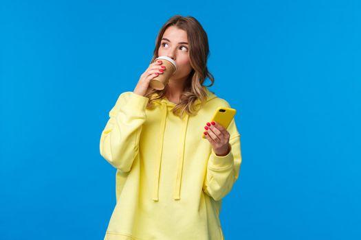 Thoughtful cute young blond female smm manager working freelance, drinking coffee from take-away cup look aside pondering and holding mobile phone, standing blue background.