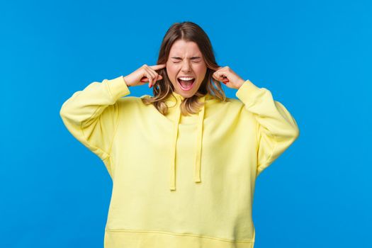 Annoyed and disturbed bothered blond woman shut ears and scream as hear loud annoying and disturbing noise, feel discomfort from living dormitory, standing blue background.