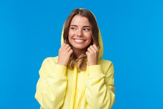 Close-up portrait beautiful charming blond girl put on hood of sweatshirt and smiling, look away, feel chilly as walking on spring day, standing carefree over blue background. Copy space