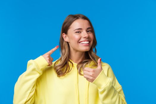 Cheerful and pleased, satisfied good-looking european female pointing at pierced ear and smiling delighted, show thumbs-up like her new jewelry, standing blue background. Copy space