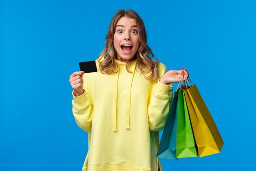 Excited happy blond girl ready waste all money on her bank account, scream thrilled and joyful during shopping, holding bags with goods and credit card, stand blue background.