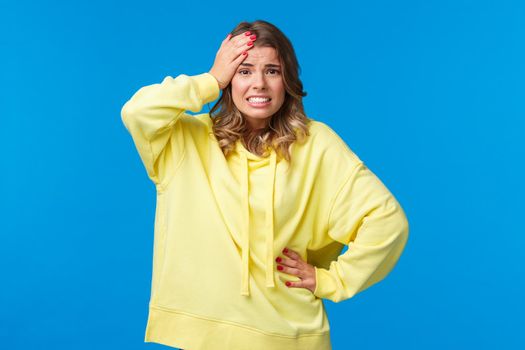 Embarrassed and worried girl bumped into car, feel anxious and concerned, touch head troubled, grimacing with awkward grin, face problematic situation, stand blue background.