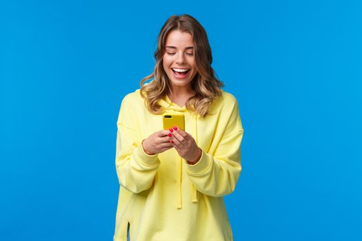 Technology and people concept. Happy smiling blond girl using mobile phone in yellow case, messaging look at smartphone display with pleased expression, send funny meme, blue background.