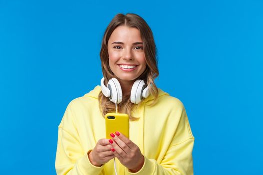 Close-up portrait of young blond european hipster girl with blond short hair and headphones over neck, holding smartphone, texting message, use mobile phone, blue background. Copy space