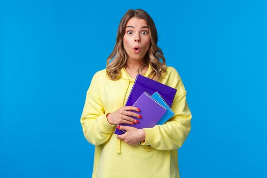 Waist-up portrait of surprised and wondered blonde caucasian female say wow and express amazement hear interesting news, gossiping in college, holding notebooks and essay, blue background.