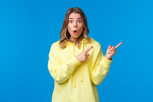 Have you seen it. Impressed and stunned blond caucasian female in yellow hoodie discuss recent events, gossips with friend, look at you and pointing right with dropped jaw, blue background.