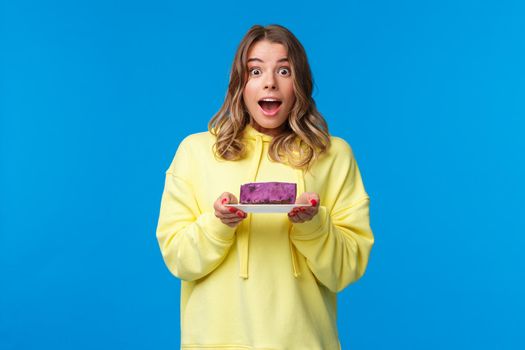Celebration, party and lifestyle concept. Excited and happy cheerful blond girl on diet eating tasty fruit cake, looking surprised and joyful camera, standing blue background.