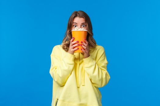 Leisure, fun and youth concept. Thrilled scared blond girl hiding behind popcorn box and stare frightened at screen as watching horror movie in cinema, express fear with eyes, blue background.