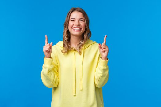 Cheerful happy smiling girl with blond hair in yellow hoodie, pointing fingers up and laughing carefree, recommend product, promo of subscribtion or company banner on blue background.