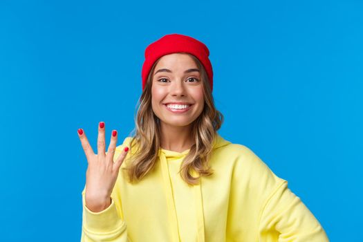 Happy smiling female student in hipster red beanie, showing number four, fourth and grinning at camera optimistic, make reservation or order, standing blue background upbeat. Copy space