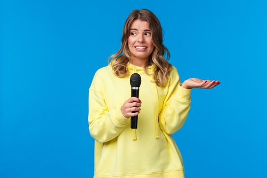 Awkward cute blond girl dont know lyrics as singing karaoke, looking at friend nervous smile, embarrassed sing in fron of people, hold microphone, standing blue background.
