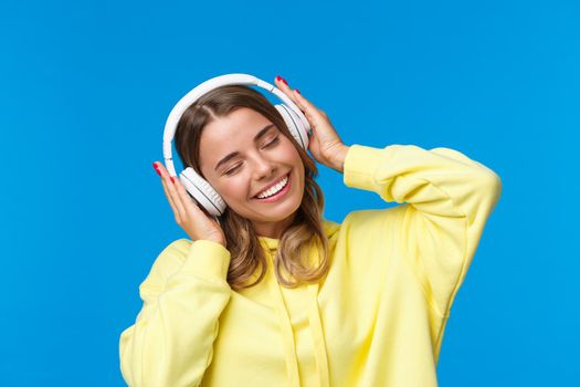 Music, lifestyle and youth concept. Close-up portrait of joyful cute young blond girl listening to favorite song in headphones, vibing, smiling delighted, standing blue background.