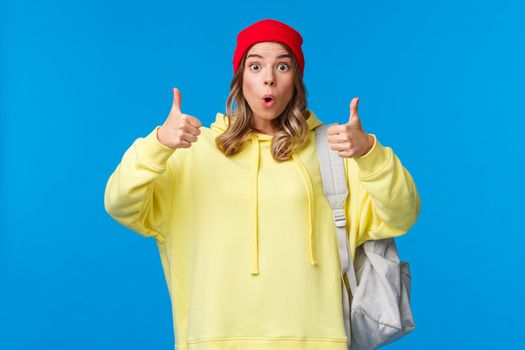 Wow really cool, good job. Impressed good-looking blond young female student in red beanie and yellow hoodie, carry backpack, praise friend nice work, show thumbs-up in approval or like.