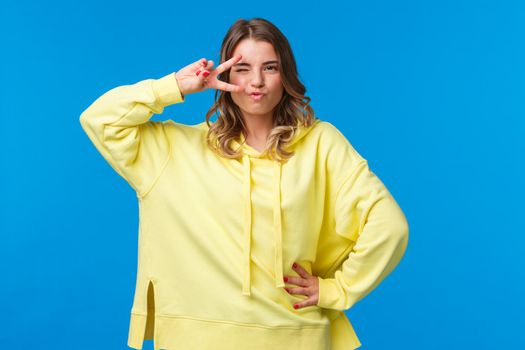 Portrait of sassy alluring blond caucasian woman dancing disco, show peace sign near eye and looking daring and sexy camera, standing in assured pose over blue background.