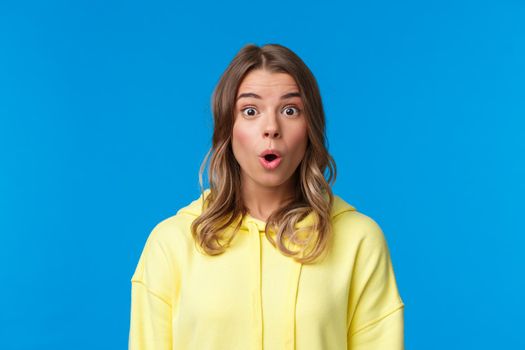 Close-up portrait of intrigued and curious good-looking young teenage girl with blond short hair, say wow, staring impressed and speechless at camera, standing blue background.