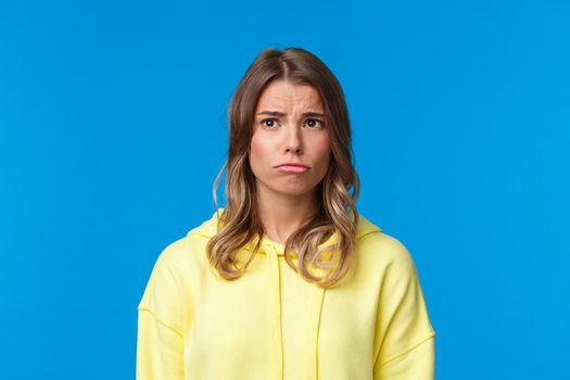 Close-up portrait gloomy sad cute jealous blond girl in yellow hoodie, pouting looking with regret upper left corner, feeling moody and uneasy, standing blue background unhappy. Copy space