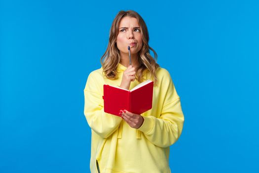 Thoughtful blond european female making plans, touching lip with pen and looking up pondering while thinking taking notes, holding red notebook, standing blue background. Copy space