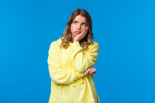 Bored and pensive young blond woman, thinking dont know what do, trying make-up plan how get rid bothring situation, standing blue background clueless look upper left corner reluctant.