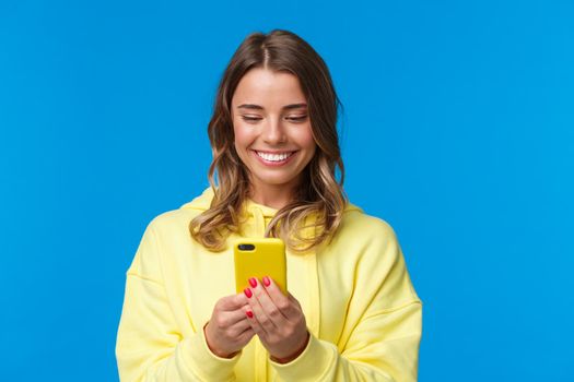 Close-up portrait silly beautiful blond european woman in yellow hoodie, smiling at mobile phone display as watching lovely video, messaging with boyfriend, standing blue background.