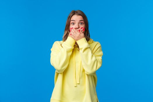 Shocked and speechless impressed blond woman close mouth with hands and stare astonished, hear shook rumour, gossiping make astounded face, standing blue background.