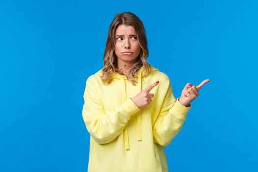 Jealous and gloomy, uneasy blond girl staring sad right, pointing fingers at something bothers her, feel regret or offended, sulking and frowning, being mocked by friends, standing blue background.