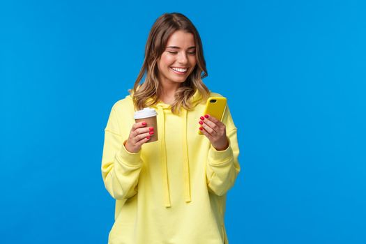 Carefree joyful european female in yellow hoodie, holding take-away coffee and browsing internet in mobile phone, texting friend laughing over funny video or meme, blue background.