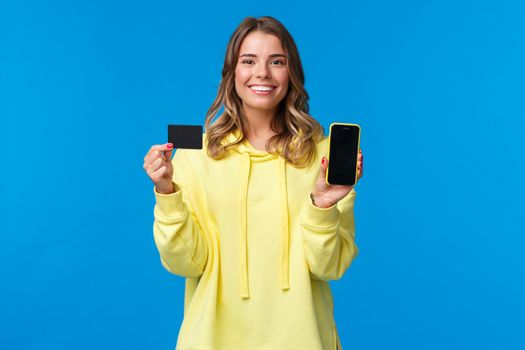 Banking and finance concept. Portrait of happy smiling blond young girl using credit card, showing it and smartphone display, satisfied with new application helps shopping online, blue background.