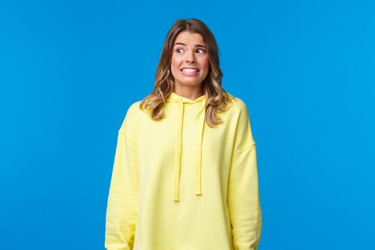 Anxious and embarrassed, alarmed insecure girl have panic and nervous thoughts of someone found out her dirty little secret, smile awkward and worried look around, stand blue background.