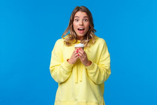 Surprised cute blond caucasian girl react to amazing news or awesome promo in her favorite cafe, holding take-away cup of coffee gasping and look camera fascinated, blue background.