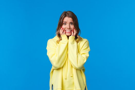 Embarrassed and afraid young timid blond caucasian female in yellow hoodie panicking, biting fingernails from fear and worries, looking scared with alarmed expression, blue background.