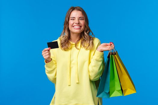 Happy smiling pretty girl using her credit card, deposit money for shopping, holding bags with clothes and grinning delighted, finally getting ready for summer vacation, blue background.