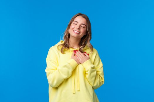 Romantic alluring blond caucasian girlfriend in yellow hoodie, press hands to heart and close eyes with lovely smile recall beautiful memory, imaging perfect date, standing blue background.