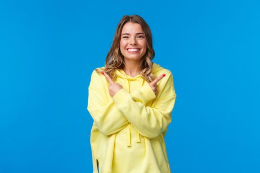 Confident pleasant, friendly-looking caucasian female showing two variants, give opportunity to choose what best, pointing fingers sideways and smiling camera, blue background.