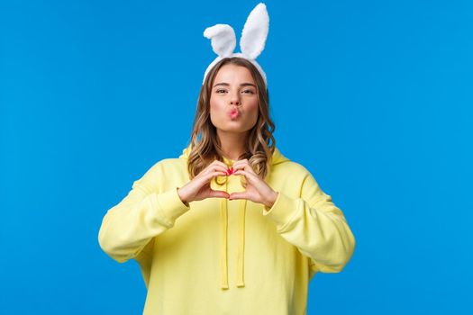 Holidays, traditions and celebration concept. Funny and cute silly blond girl in yellow hoodie and rabbit ears showing heart gesture, make mwah kiss expression, spread love and positivity.