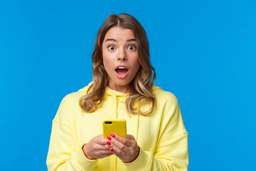 Close-up portrait entertained and surprised cute blond girl look with disbelief and amazement camera after reading interesting news online, holding mobile phone, standing blue background.