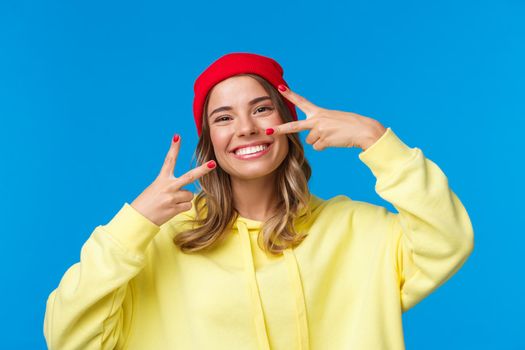 Carefree good-looking european female in red beanie and yellow hoodie, smiling kawaii show peace gestures as sending positive vibes, standing upbeat over blue background.