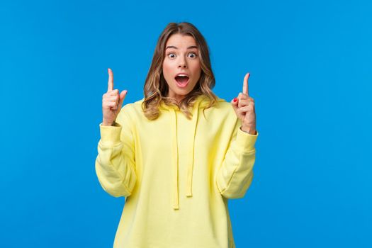 Speechless impressed young caucasian female staring at camera with dropped jaw, gasping astonished pointing fingers up, talking about awesome promo offer discount, stand blue background.
