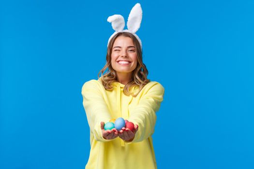 Holidays, people and emotions concept. Tender cute blond caucasian female giving Easter eggs to you with beaming pleasant smile, wearing rabbit ears, celebrating religious holiday.