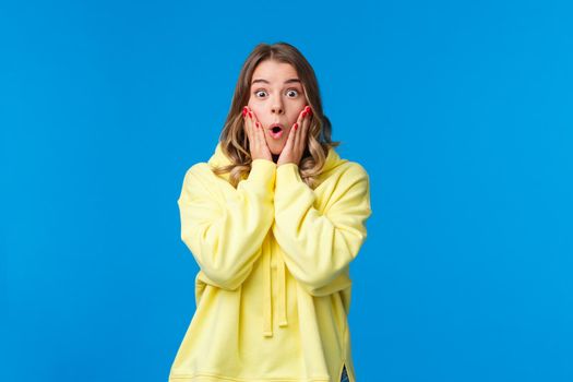 Impressed speechless young blond woman open mouth stare startled camera and say wow, touch cheeks from amazement stare disbelief and amazement, hear fantastic news, blue background.