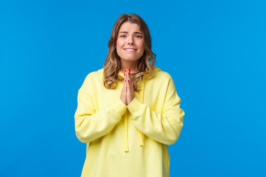 Cute girl saying please and asking you favour, need something asap, hold hands in pray smiling and looking hopeful camera as begging, pleading to lend money, blue background.