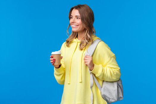 Carefree pretty young female student drinking coffee from paper cup while heading university, holding backpack and looking aside with pleased relaxed smile, lifestyle and people concept.