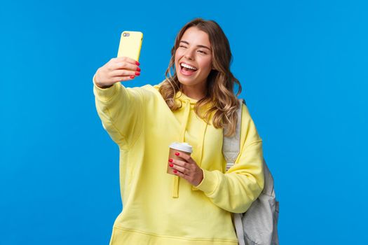 Lifestyle, university and education concept. Cheerful good-looking blond female student or traveller taking selfie with backpack and take-away coffee in paper cup, wink to mobile phone camera.