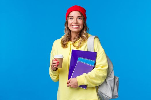 Happy excited cute girl starting senior year in college, holding notebooks and take-away coffee, carry backpack, smiling upbeat camera, wearing hipster beanie and yellow hoodie.