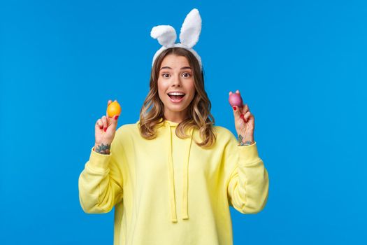 Holidays, traditions and celebration concept. Cute blond caucasian sister want play game with brother holding two painted Easter eggs and smiling, wearing rabbit ears, blue background.