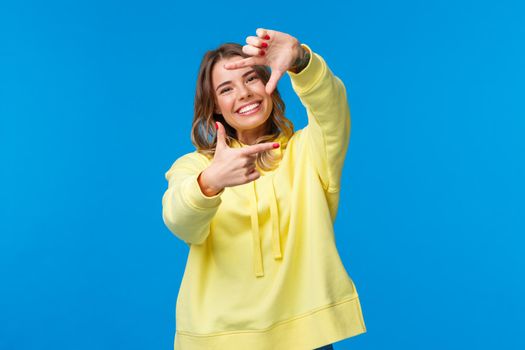Inspiration, lifestyle and people concept. Portrait of cheerful creative blond girl searching perfect angle for shot, look at camera through frame gesture and smiling, blue background.