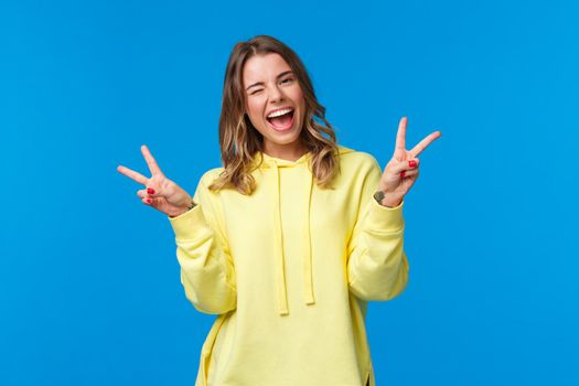 Lets have some fun. Carefree upbeat friendly-looking kawaii blond girl in yellow hoodie, wink and smiling white teeth as showing cute peace gestures, standing blue background.