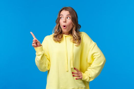Thrilled speechless good-looking blonde female in yellow hoodie react to something shocking and interesting, pointing finger and looking left saying wow, gossiping, stand blue background.