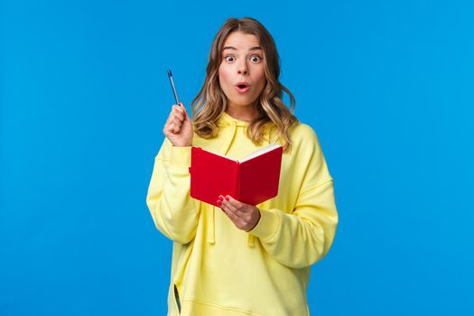 Girl doing homework finally have great plan, made-up idea for story as writing it in red notebook, raise pen eureka gesture, looking amazed camera, standing blue background in yellow hoodie.