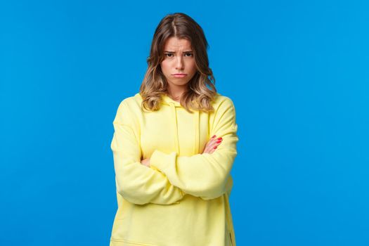 Gloomy and moody cute blond teenage girl look from under forehead and sulking at camera, feeling offended or upset, cross hands chest insulted waiting apology, blue background.